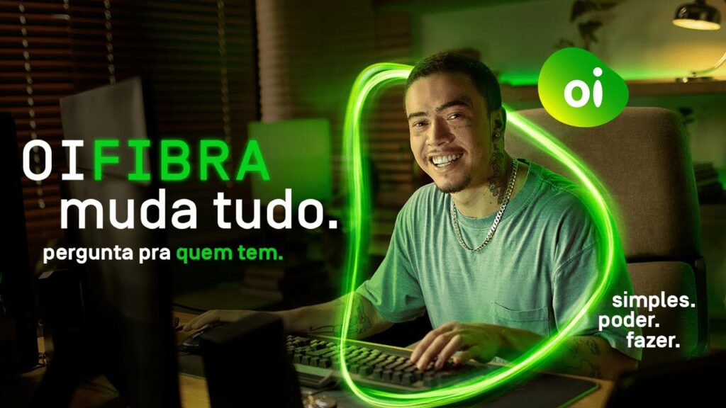 A imagem é uma campanha da Oi Fibra com o influencer Whindersson Nunes, que se encontra mexendo em um computador e sorrindo para a foto.