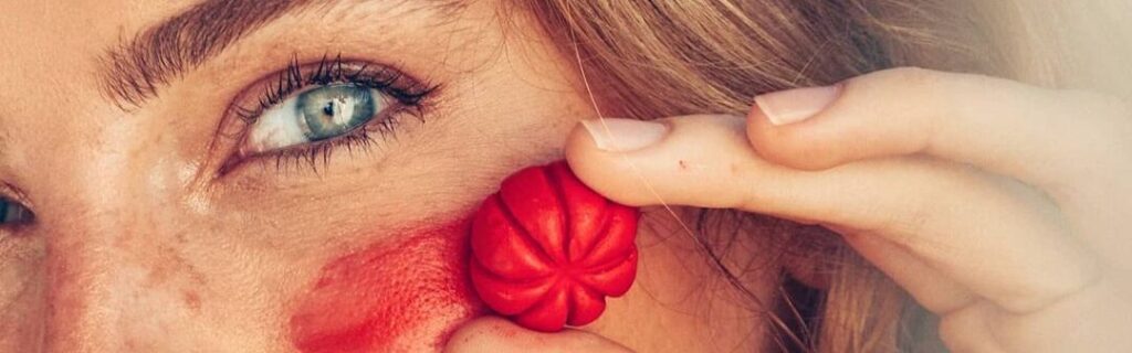 Imagem promocional da marca Amo Karité, mostrando uma modelo aplicando maquiagem sólida vermelha como blush.